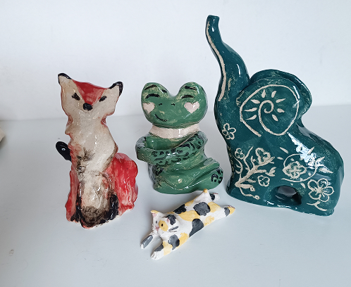 Cuatro figuras de cerámica hechas a mano: un zorro rojo, una rana verde sentada, un gato calicó tumbado y un elefante verde con grabados florales.