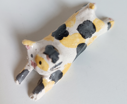 Figura de cerámica de un gato calicó estirándose, con manchas negras y amarillas sobre fondo blanco.