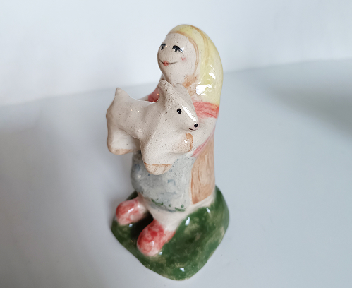 Pequeña escultura de cerámica pintada a mano que representa a una mujer sosteniendo una oveja blanca.