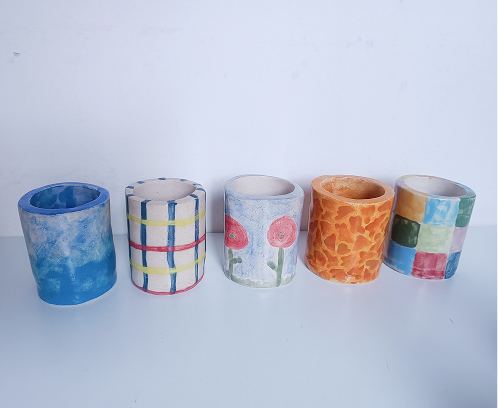 Fila de cinco botes de cerámica hechos a mano con diferentes diseños: degradado azul, rayas, flores rojas, manchas naranjas y cuadros de colores.
