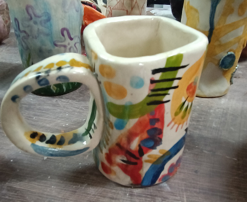Taza de cerámica artesanal de forma orgánica, decorada con motivos abstractos vibrantes en azul, amarillo, verde y rojo sobre un fondo blanco brillante.