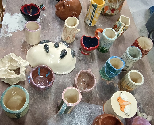 Vista aérea de una mesa de trabajo con múltiples piezas de cerámica: tazas con esmaltes de colores, pequeños cuencos y una pieza central blanca con relieves oscuros.