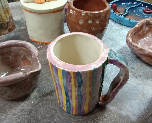 Vista aérea de una mesa de trabajo con múltiples piezas de cerámica: tazas con esmaltes de colores, pequeños cuencos y una pieza central blanca con relieves oscuros.