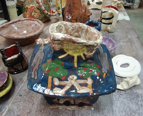 Vista aérea de una mesa de trabajo con múltiples piezas de cerámica: tazas con esmaltes de colores, pequeños cuencos y una pieza central blanca con relieves oscuros.
