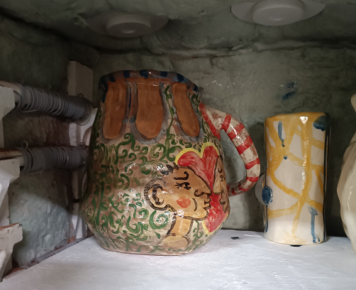 Jarra de cerámica artesanal con forma irregular, decorada con ilustraciones de rostros de perfil en tonos tierra y un patrón de volutas verdes sobre fondo crema.