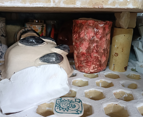 Detalle de estantería de secado en un taller de cerámica, mostrando piezas en proceso, una taza con textura rojiza y herramientas de soporte refractario.