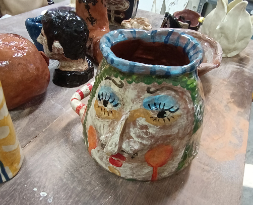 Primer plano de una jarra cerámica con volumen, destacando un rostro esculpido y pintado con grandes ojos azules, mejillas anaranjadas y un asa con rayas rojas y blancas.