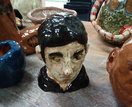 Escultura cerámica de una cabeza masculina con acabado brillante, cabello negro y expresión introspectiva, rodeada de otras piezas en un taller.